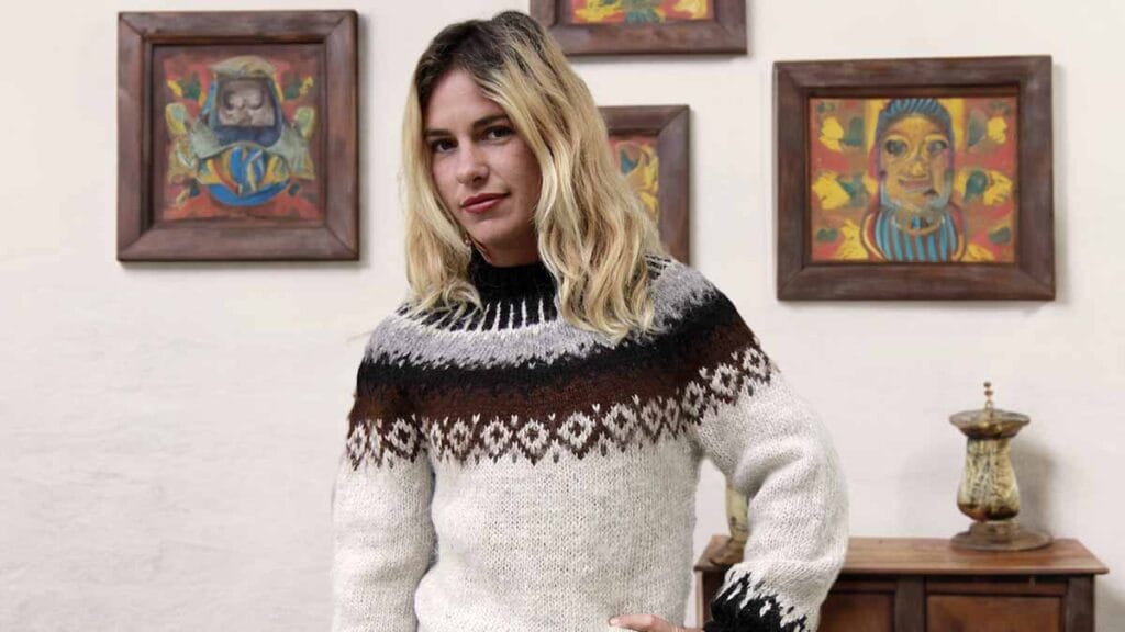 A blonde woman wears a handmade alpaca wool sweater. Natural white sweater with geometric design on the shoulders.